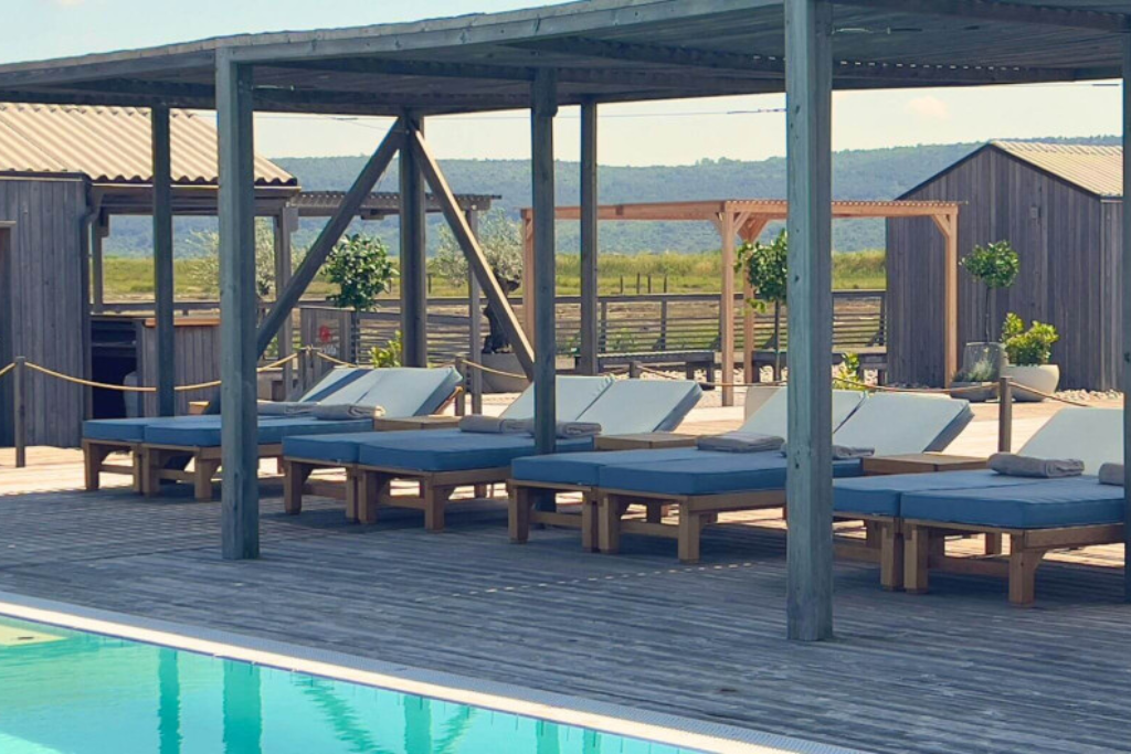 Poolside deck with blue-cushioned loungers under a wooden pergola and towels at the end of each bed in a sunny outdoor area