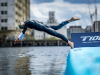 Triathlete in a wetsuit diving from a blue ramp into the water at a race start, with a T100 Triathlon World Tour banner nearby.
