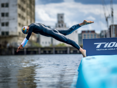 Triathlete in a wetsuit diving from a blue ramp into the water at a race start, with a T100 Triathlon World Tour banner nearby.