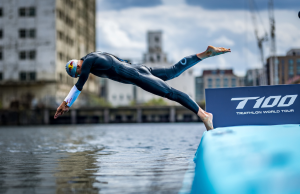 Triathlete in a wetsuit diving from a blue ramp into the water at a race start, with a T100 Triathlon World Tour banner nearby.