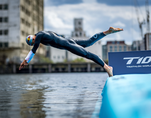 Triathlete in a wetsuit diving from a blue ramp into the water at a race start, with a T100 Triathlon World Tour banner nearby.