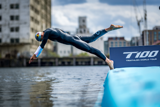 Triathlete in a wetsuit diving from a blue ramp into the water at a race start, with a T100 Triathlon World Tour banner nearby.