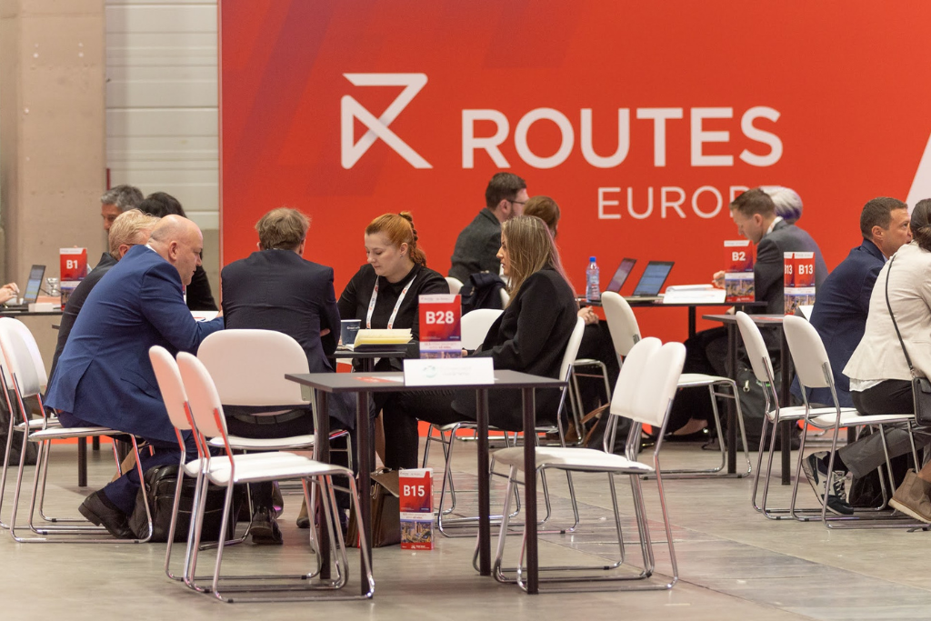 Groups of professionals in business attire sit around round tables in a conference hall, with a bright orange ROUTES EUROPE backdrop in the background.
