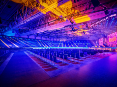 Empty arena with rows of chairs facing a stage with large LED screens and colorful blue, purple, and yellow lighting.