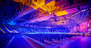 Empty arena with rows of chairs facing a stage with large LED screens and colorful blue, purple, and yellow lighting.