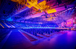 Empty arena with rows of chairs facing a stage with large LED screens and colorful blue, purple, and yellow lighting.