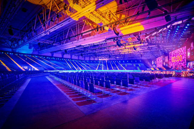 Empty arena with rows of chairs facing a stage with large LED screens and colorful blue, purple, and yellow lighting.