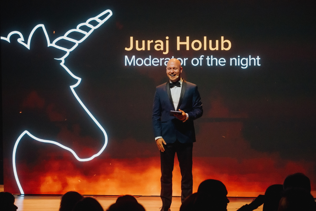 Male host in a navy tuxedo on stage, holding a card, with neon unicorn outline and the text 'Juraj Holub Moderator of the night' on screen.
