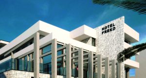 Modern white hotel with stone accent wall and large glass windows under a blue sky; sign reads HOTEL PREKO