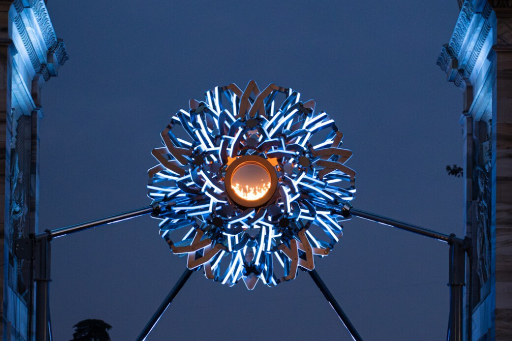 balich_wonder_studio Blue neon dandelion-like sculpture with an orange core, mounted on a metal tripod between stone arches at night.