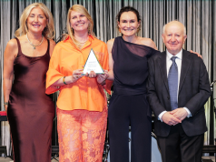 Four adults in formal attire pose on a stage; the woman in orange holds a triangular award while the others smile beside her.
