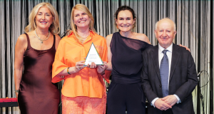 Four adults in formal attire pose on a stage; the woman in orange holds a triangular award while the others smile beside her.