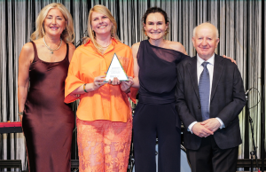 Four adults in formal attire pose on a stage; the woman in orange holds a triangular award while the others smile beside her.