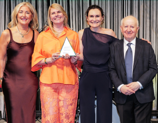 Four adults in formal attire pose on a stage; the woman in orange holds a triangular award while the others smile beside her.