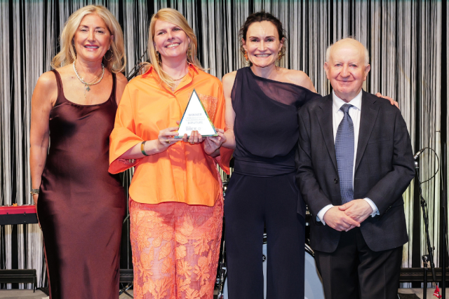 Four adults in formal attire pose on a stage; the woman in orange holds a triangular award while the others smile beside her.