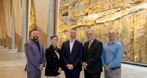 Five professionals posing for a group photo in a bright, modern interior with a rock wall backdrop.