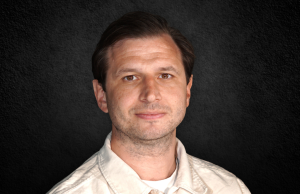 Portrait of a man with short dark hair wearing a light beige shirt, against a dark textured background.
