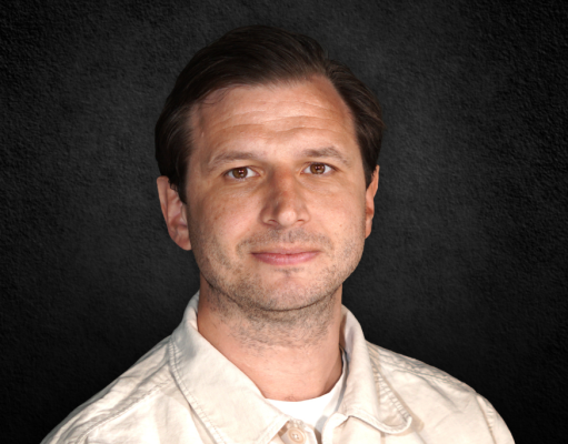 Portrait of a man with short dark hair wearing a light beige shirt, against a dark textured background.