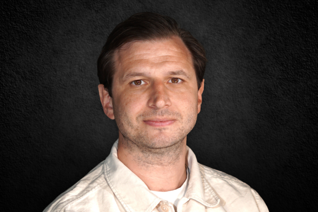 Portrait of a man with short dark hair wearing a light beige shirt, against a dark textured background.