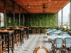 Rooftop cafe seating area with tall wooden tables and stools on the left, blue-patterned chairs and white tables on the right, glass walls, and a green leafy wall at the back with city buildings in the distance.