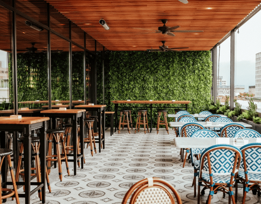 Rooftop cafe seating area with tall wooden tables and stools on the left, blue-patterned chairs and white tables on the right, glass walls, and a green leafy wall at the back with city buildings in the distance.