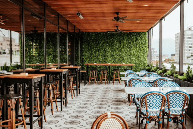 Rooftop cafe seating area with tall wooden tables and stools on the left, blue-patterned chairs and white tables on the right, glass walls, and a green leafy wall at the back with city buildings in the distance.