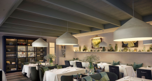 Elegant restaurant dining room with white-tablecloth tables, green upholstered chairs, and large pendant lights, creating a warm, upscale ambiance.