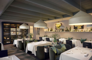 Elegant restaurant dining room with white-tablecloth tables, green upholstered chairs, and large pendant lights, creating a warm, upscale ambiance.