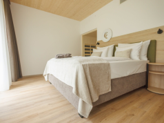Bright hotel bedroom with a king bed, white linens, beige throw, and a curved wood nightstand by the headboard.