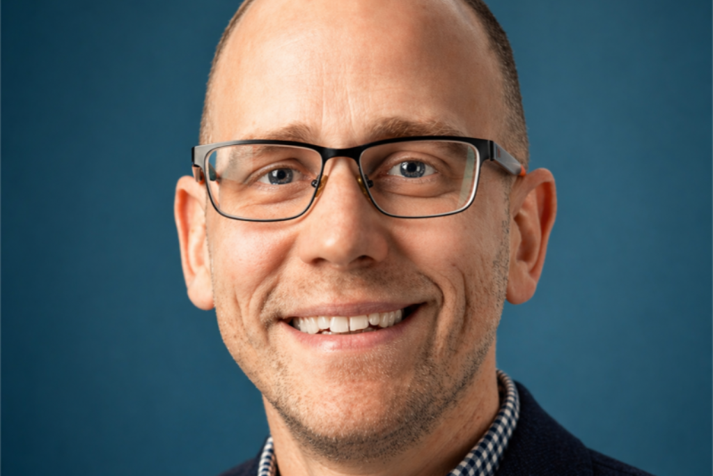 christian_skelton Close-up portrait of a smiling man wearing rectangular glasses against a blue background.