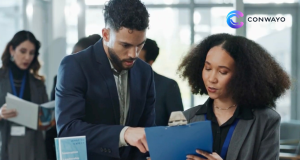 Two professionals review a blue clipboard at a conference table, with a woman in a gray blazer and a man in a navy suit, logo visible in the top-right corner.