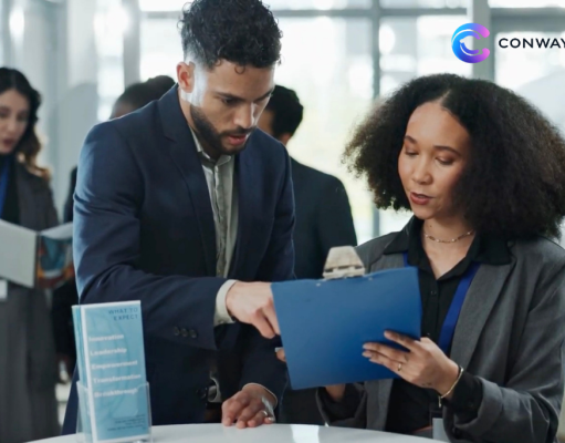 Two professionals review a blue clipboard at a conference table, with a woman in a gray blazer and a man in a navy suit, logo visible in the top-right corner.