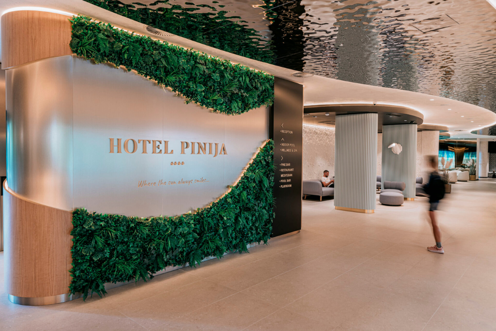 hotel_pinija Hotel Pinja lobby entrance with a curved green plant wall and a sleek metal sign reading 'HOTEL PINJA' near the reception corridor. A person walks through the spacious, modern lobby.