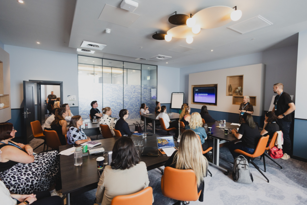 mi_healthcare_brighton Group of people seated around tables in a modern conference room, listening to a presenter at the front near a screen.
