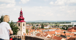 Woman in a white shirt with a red bicycle, looking over a town with orange-red rooftops and a tall red-domed church tower.