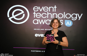 Smiling woman in a black dress on stage holds a pink-and-black award against a dark backdrop that reads 'event technology awards'.
