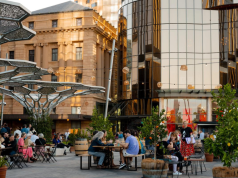 Adelaide in global top three for business travel growth People sit at wooden tables in an outdoor plaza with potted trees, dining and chatting, beside modern glass buildings and historic architecture.