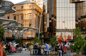 Adelaide in global top three for business travel growth People sit at wooden tables in an outdoor plaza with potted trees, dining and chatting, beside modern glass buildings and historic architecture.