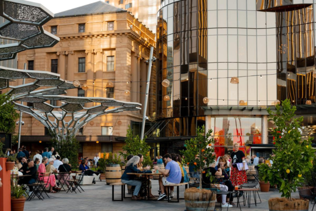People sit at wooden tables in an outdoor plaza with potted trees, dining and chatting, beside modern glass buildings and historic architecture.