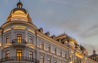 Elegant neoclassical hotel facade at dusk with illuminated 'CORINTHIA' signs on domed towers and ornate balconies