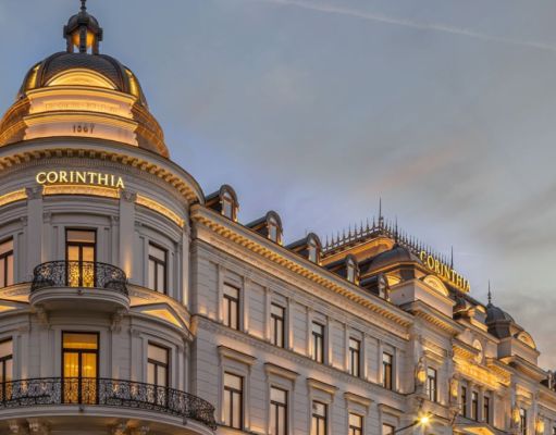 Elegant neoclassical hotel facade at dusk with illuminated 'CORINTHIA' signs on domed towers and ornate balconies