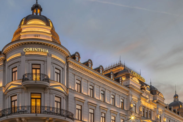 Elegant neoclassical hotel facade at dusk with illuminated 'CORINTHIA' signs on domed towers and ornate balconies