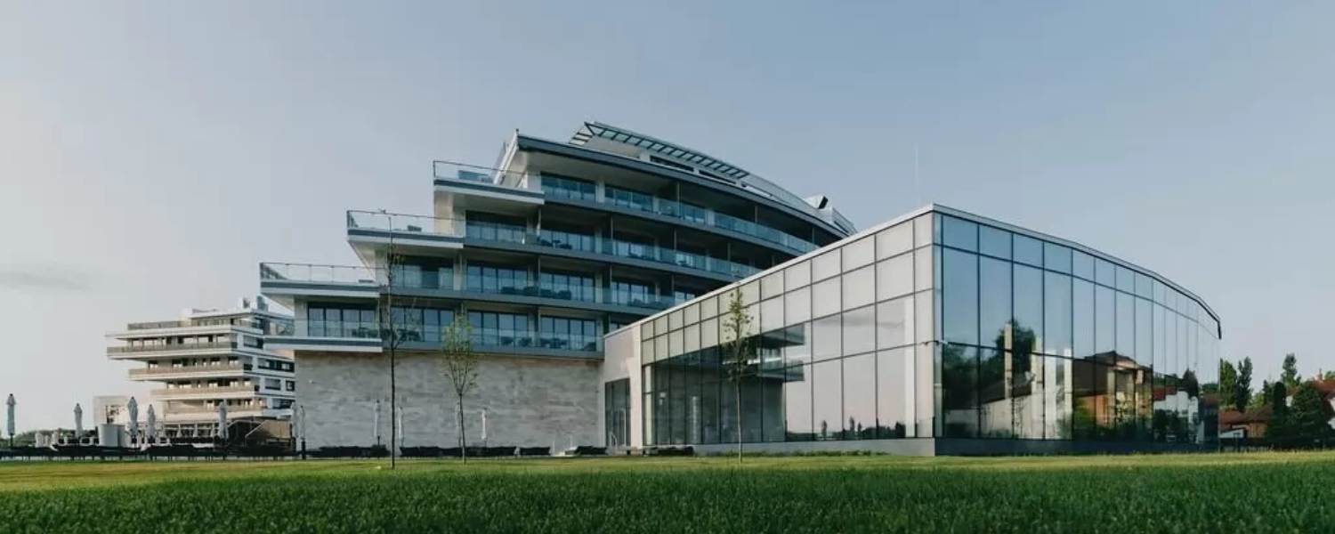 Modern glass office building with curved facade and balconies on a green lawn under a clear sky.